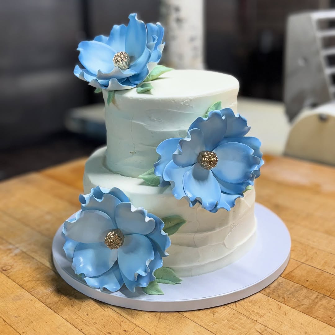 A two-tier cake with blue sugar flowers on the wooden decorating bench, the bakery workroom and mixer behind it