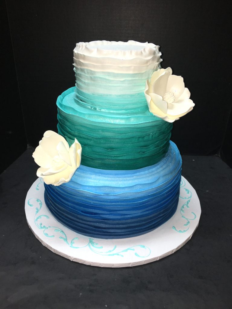 Three-tier ruffled buttercream cake shading from white to teal to navy, with two white sugar magnolias