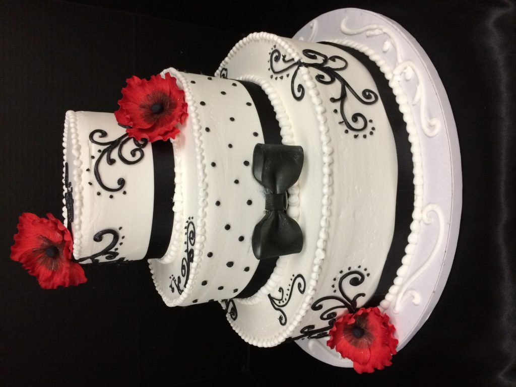 Three-tier white cake with black ribbon, black scrollwork, a black bow and red sugar poppies