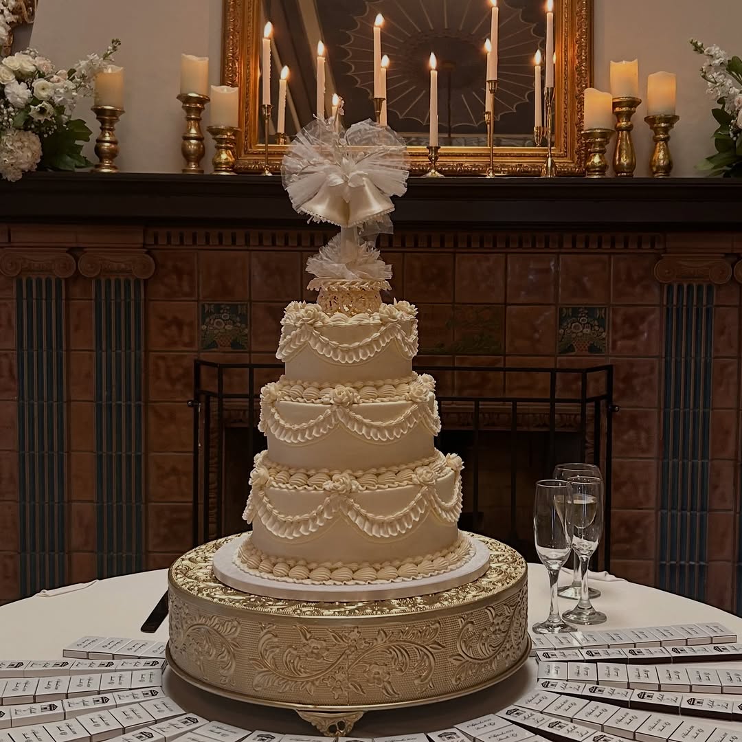 Ivory four-tier wedding cake with piped swags on a gold stand at a candlelit reception